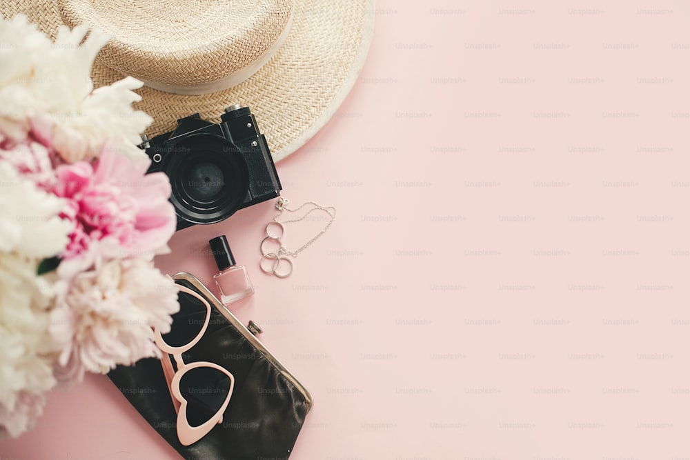 Stylish photo camera, sunglasses, jewelry,straw hat, pink and white peonies bouquet on pink paper flat lay with space for text. Hello summer.  International womens day.