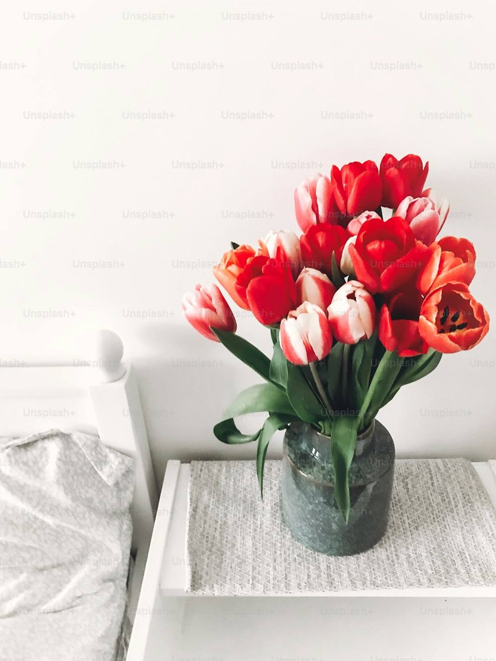 Hermosos tulipanes rojos y rosas en jarrón en mesita de noche de madera blanca cerca de una elegante cama blanca, espacio de copia. Hola concepto de primavera. Feliz día de la madre