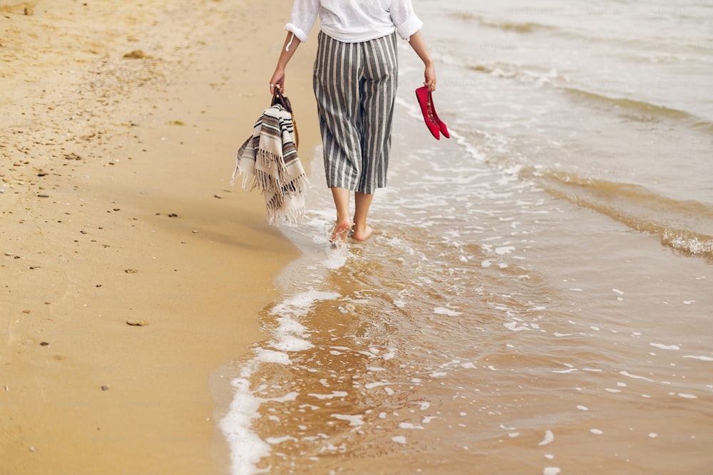 Mujer caminando descalza en la playa, vista trasera de las piernas. Joven relajándose en la playa de arena, caminando con zapatos y bolsa en las manos. Concepto de vacaciones de verano