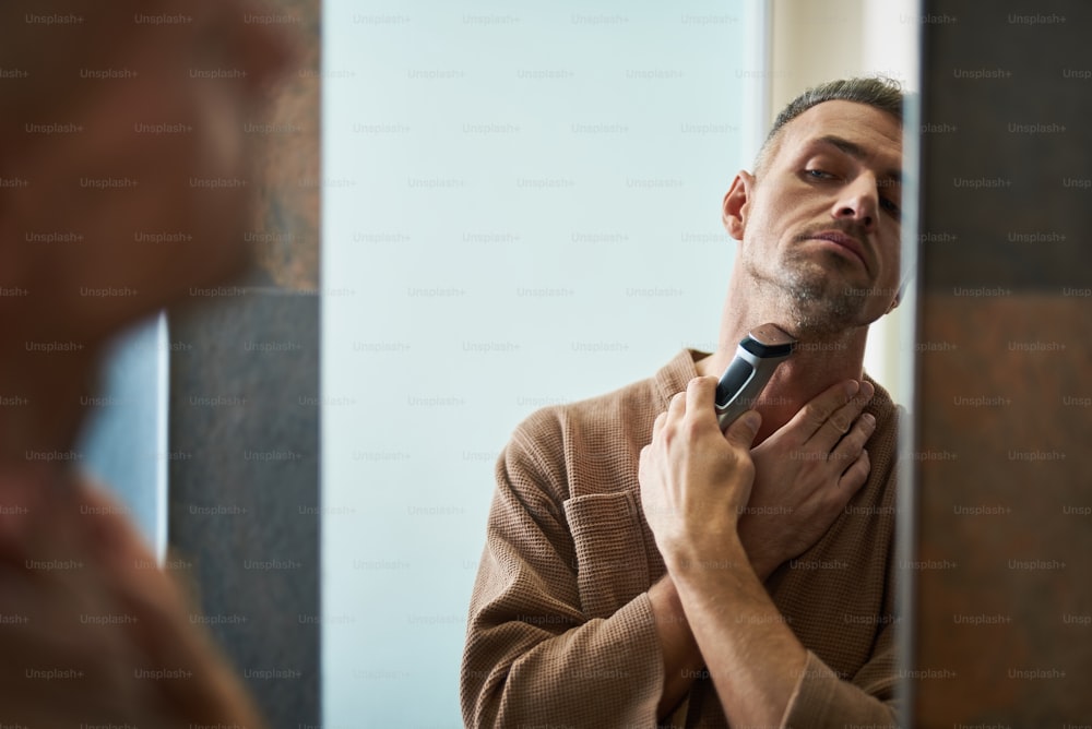 Attractive gentleman in bathrobe removing stubble from neck with shaving machine while looking in the mirror