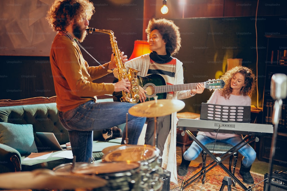 Mixed race woman playing acoustic guitar while man playing saxophone. Home studio interior. In background woman playing clavier.