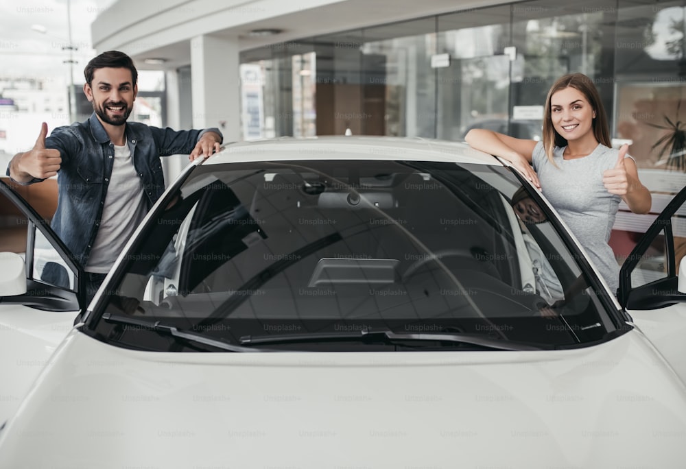 Happy beautiful couple is choosing a new car at dealership, smiling, looking at the camera and showing thumbs up.