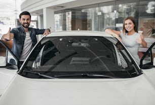 Feliz casal bonito está escolhendo um carro novo na concessionária, sorrindo, olhando para a câmera e mostrando polegares para cima.