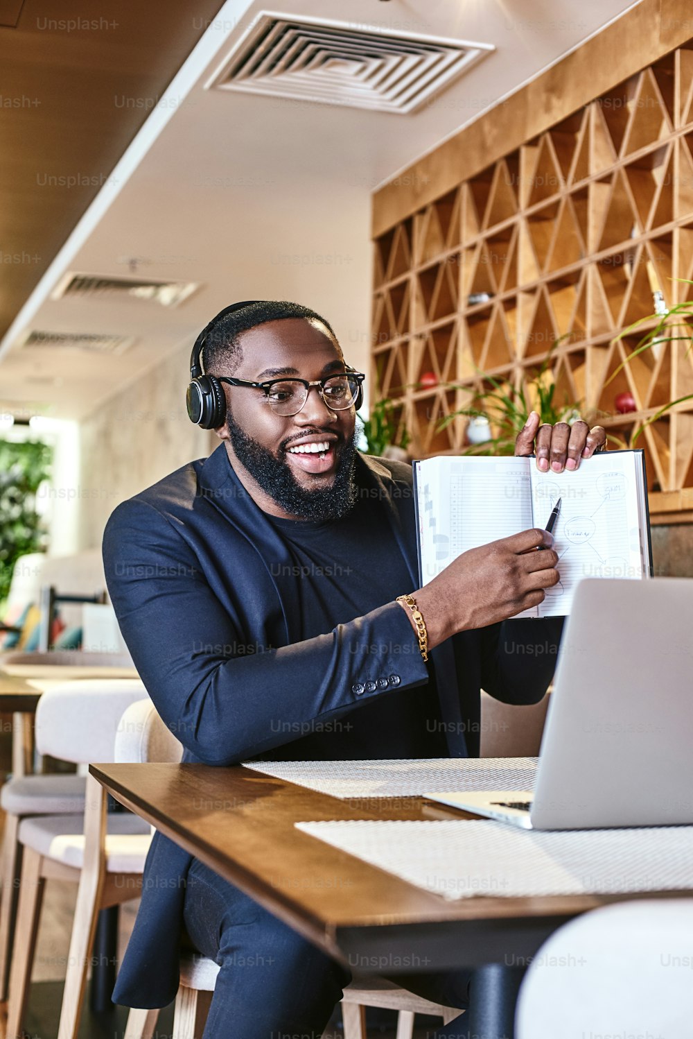 Joven hombre de negocios afroamericano con estilo positivo con chaqueta azul oscuro y anteojos sentado a la mesa, en el interior, usando auriculares inalámbricos, charlando con su jefe a través de una llamada de videoconferencia en una computadora portátil genérica, señalando el esquema dibujado en su cuaderno, mirando la pantalla y sonriendo alegremente