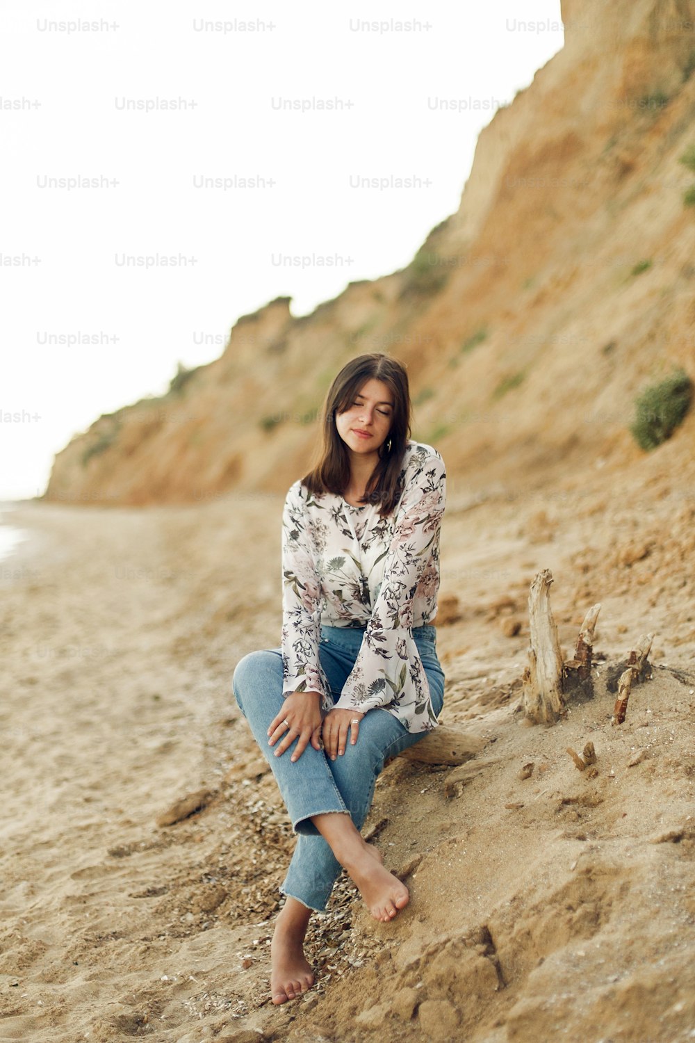 Mujer boho joven feliz sentada y relajándose en la playa soleada con roca. Chica hipster bohemia despreocupada sonriendo en el mar. Concepto de vacaciones de verano. Copiar espacio. Momento de calma