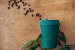 Elegante tazza da caffè ecologica riutilizzabile su sfondo in legno con chicchi di caffè e foglie di bambù verde. Vietare la plastica monouso. Concetto di zero rifiuti, posa piatta. Stile di vita sostenibile. Tazza in bambù naturale