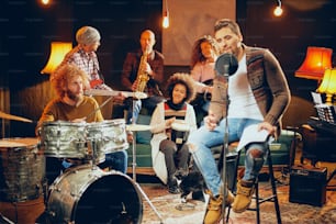 Homme chantant et assis sur une chaise pendant que son groupe joue des instruments en arrière-plan. Int�érieur du home studio.