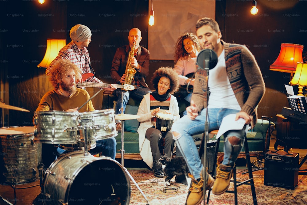 Man singing and sitting on chair while his band playing instruments in background. Home studio interior.
