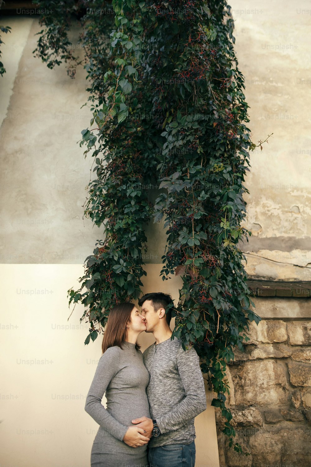 Stylish pregnant couple holding hands on baby bump, kissing under green leaves in city street. Happy young parents, mom and dad, hugging belly and embracing. Parenthood concept. True happiness