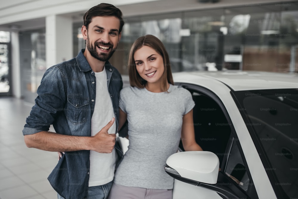 Happy beautiful couple is choosing a new car at dealership, smiling and looking at camera.
