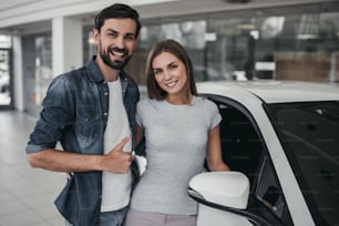 Happy beautiful couple is choosing a new car at dealership, smiling and looking at camera.