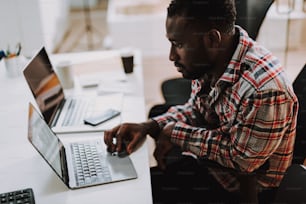 Calm young Afro-American man sitting at the white table in his office and looking at the screen of his laptop while working