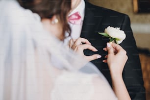 Gorgeous bride in amazing gown putting on white peony boutonniere groom suit. Beautiful woman getting ready in the morning. Wedding preparations