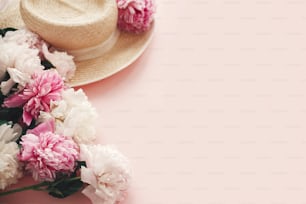 Stylish  straw rustic hat with pink and white peonies on pink paper with space for text. Hello summer. International womens day. Happy mothers day. Greeting card mockup.