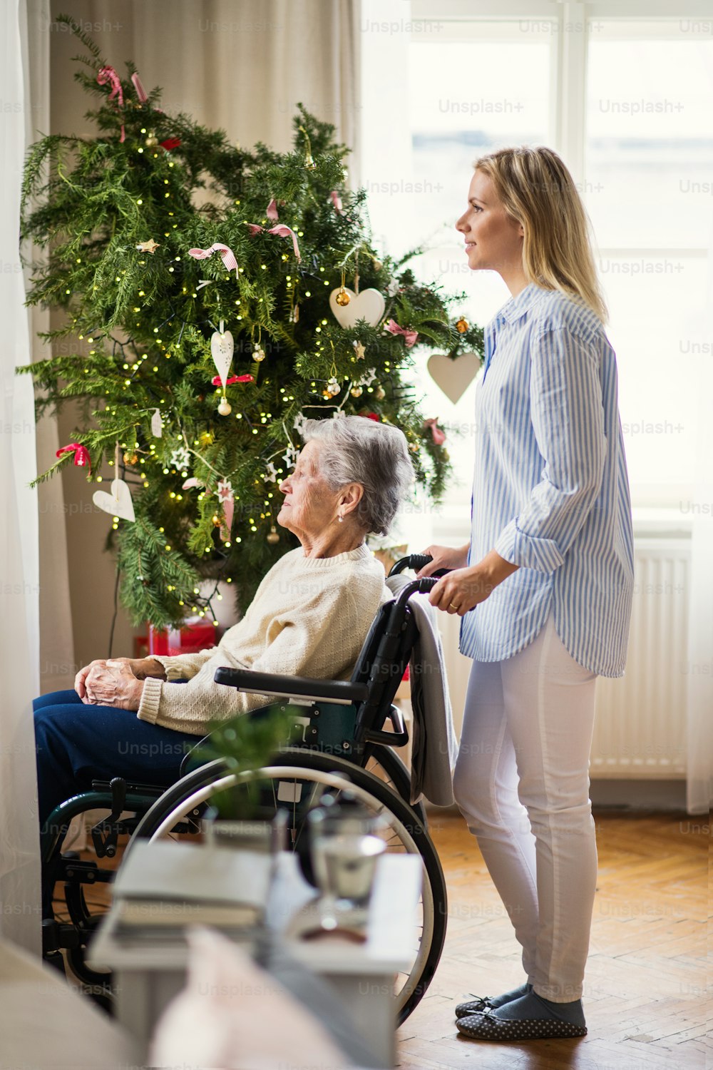 Une femme âgée en fauteuil roulant avec un visiteur de santé à la maison au moment de Noël, regardant par une fenêtre.