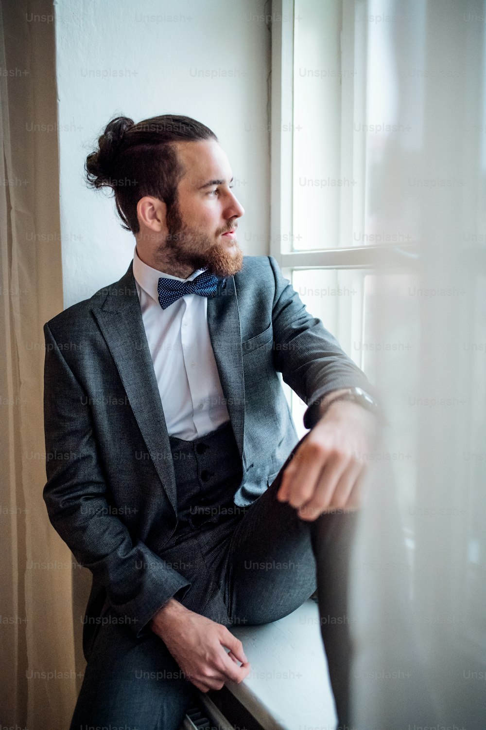 A handsome hipster young man with formal suit sitting on a window sill on an indoor party, looking out.