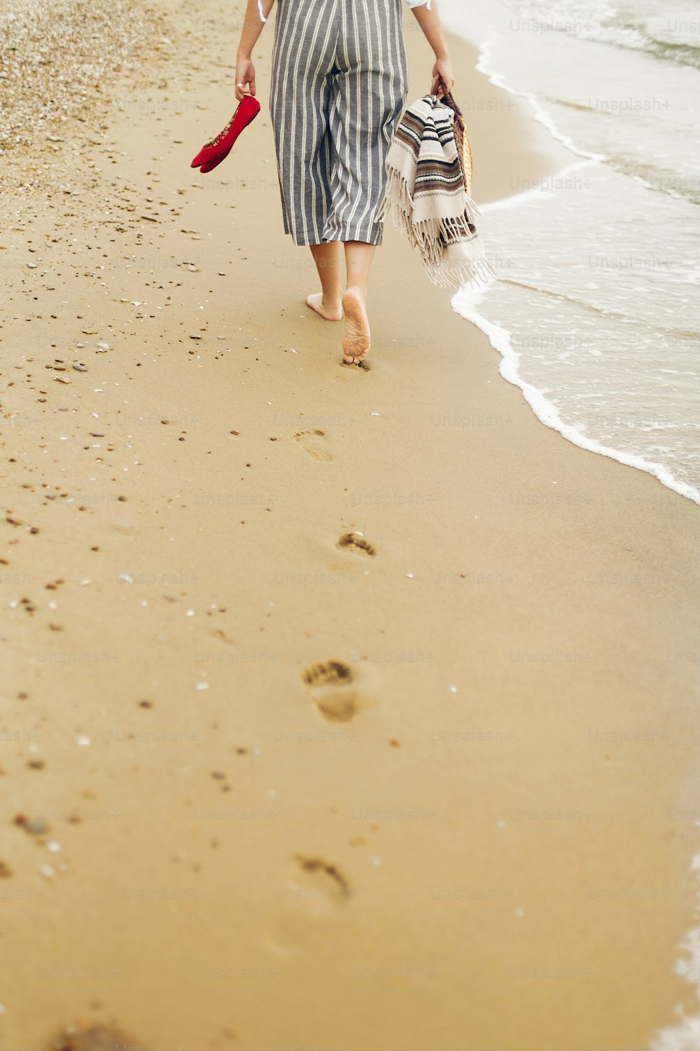 Donna che cammina a piedi nudi sulla spiaggia, vista posteriore delle gambe. Ragazza che si rilassa sulla spiaggia sabbiosa, camminando con le scarpe e la borsa in mano. Concetto di vacanza estiva