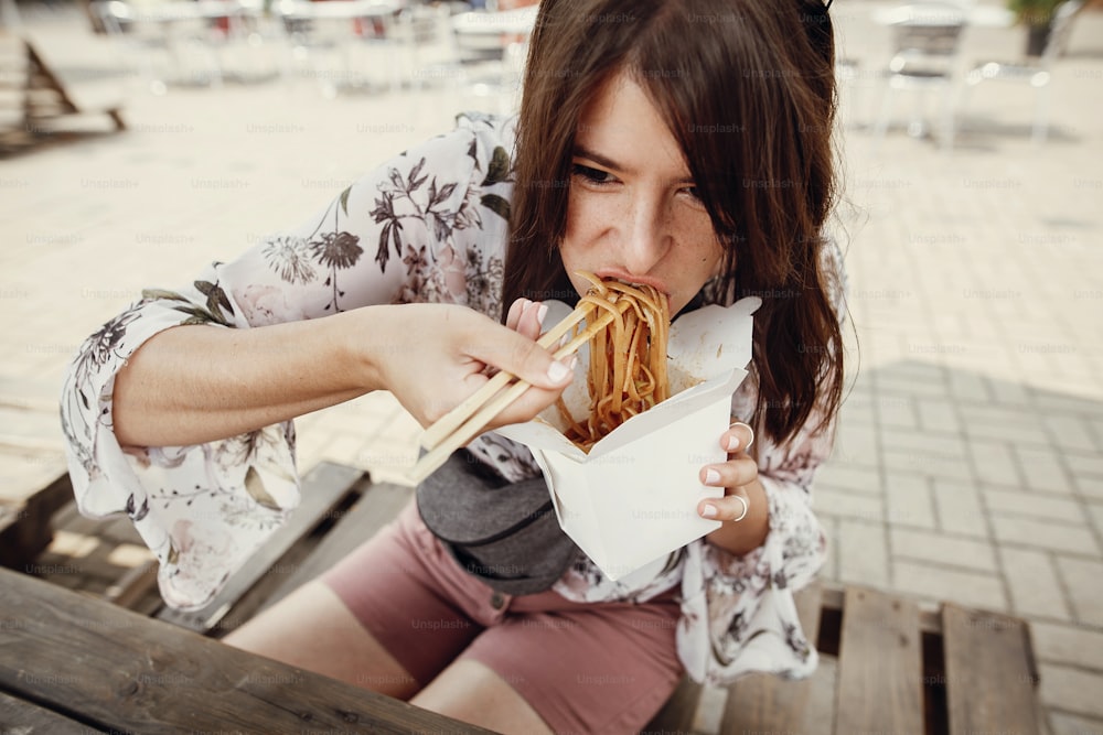 Hungry boho woman eating noodles in takeaway paper box. Food delivery. Asian Street food festival. Stylish hipster girl eating wok noodles with vegetables from carton box with bamboo chopsticks