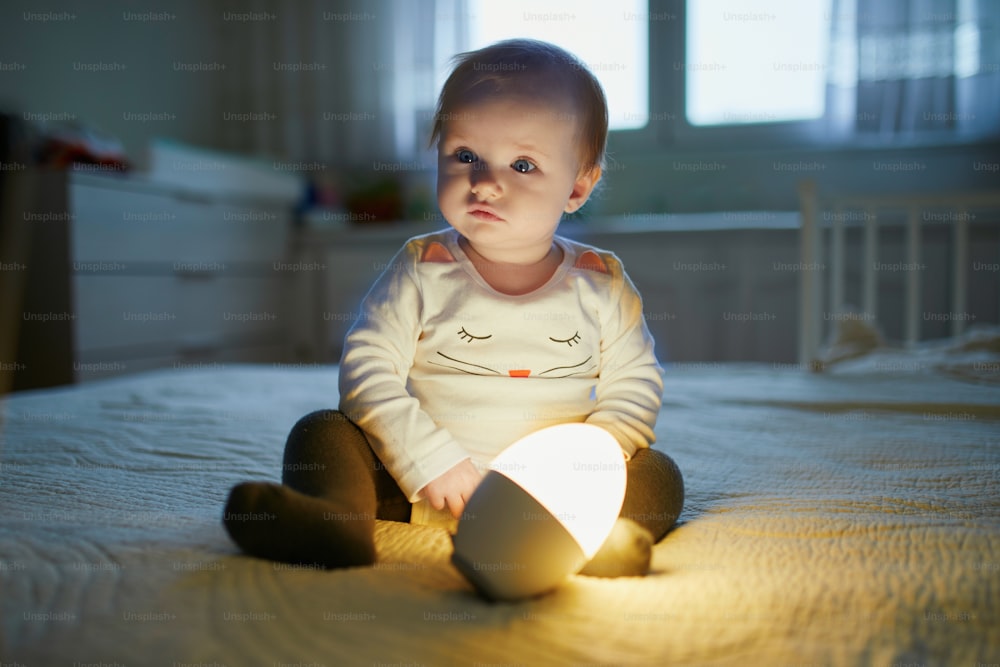 Adorable baby girl playing with bedside lamp in nursery. Happy kid sitting on bed with nightlight. Little child at home in the evening before sleep