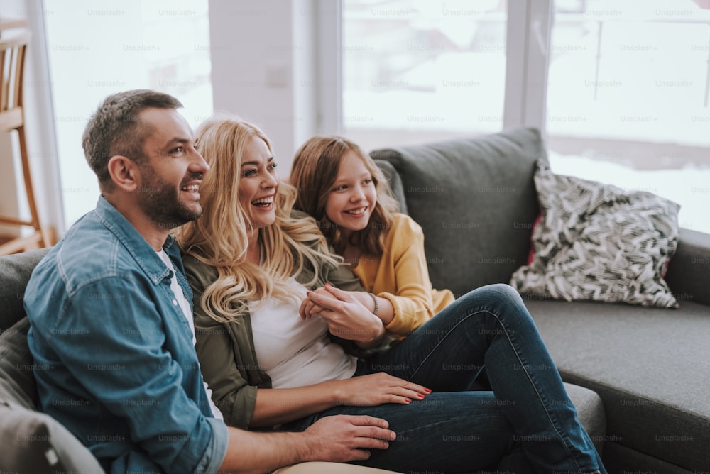 Side view portrait of handsome bearded man spending time with his beautiful wife and daughter. They watching TV and laughing