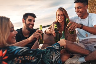 Group of young friends enjoying a day at the lake. They sitting on pier talking, laughing and drinking beers.