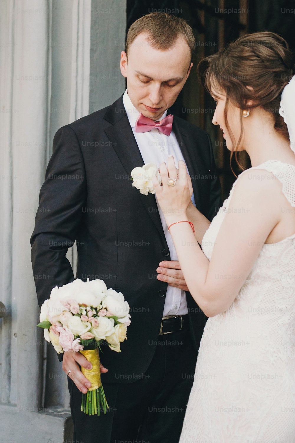 Happy Bride putting on stylish boutonniere on groom suit at doors after wedding ceremony in church. Happy stylish wedding couple during holy matrimony