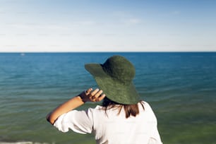 Stylish girl in hat standing on beach cliff and looking at sea in sunny evening. Happy young boho woman in hat relaxing on shore at sunset. Summer vacation. Copy space. Back view