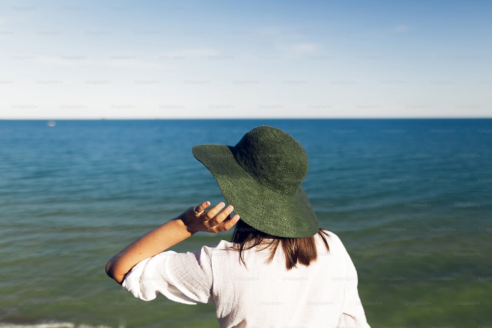 Menina elegante no chapéu de pé no penhasco da praia e olhando para o mar na noite ensolarada. Mulher boho jovem feliz no chapéu relaxando na costa ao pôr do sol. Férias de verão. Espaço de cópia. Vista traseira