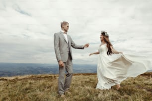 happy gorgeous bride and groom holding hands and dancing  in sun light, boho wedding couple, luxury ceremony at mountains with amazing view