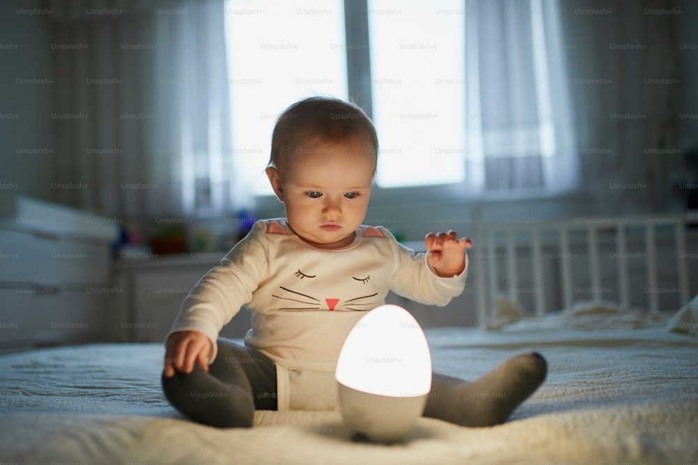 Adorable baby girl playing with bedside lamp in nursery. Happy kid sitting on bed with nightlight. Little child at home in the evening before sleep