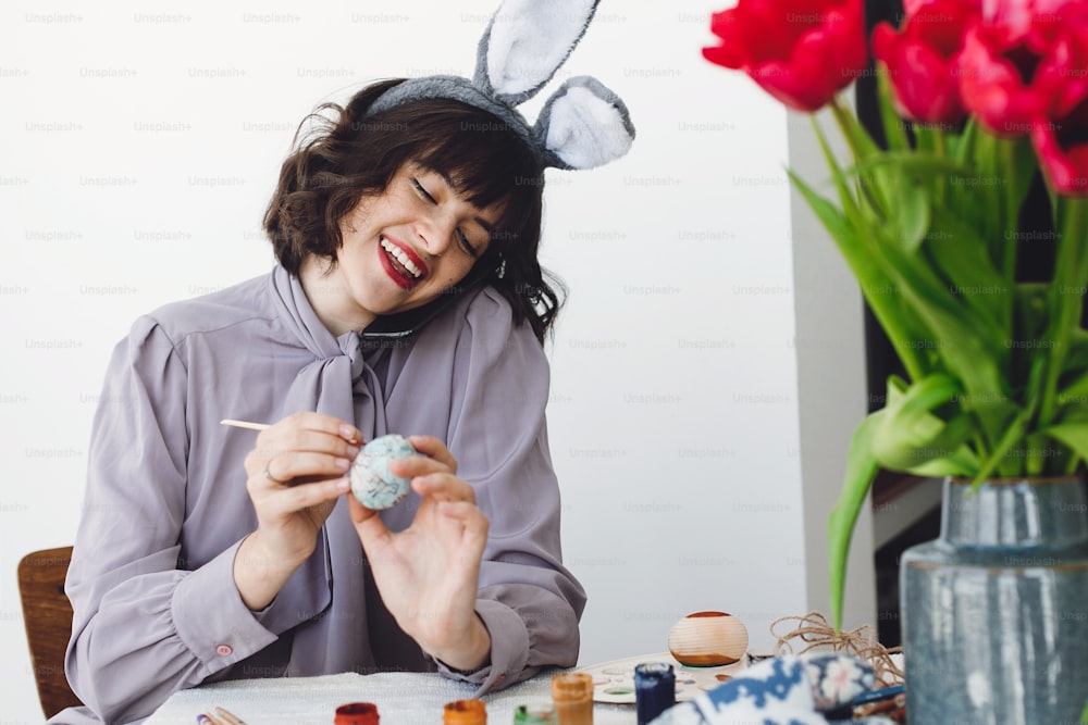 Beautiful girl in bunny ears talking on phone and smiling, painting easter egg at table with tulips in vase. Stylish young brunette woman decorating easter egg, holiday preparations. Happy Easter