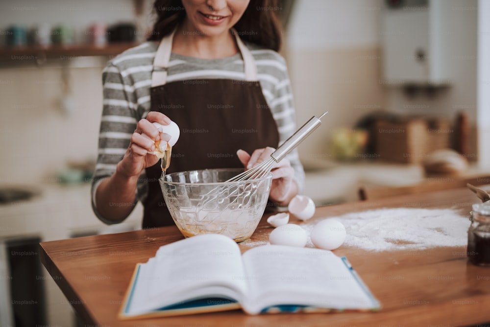 Smiling young woman is standing at table and cooking homemade pastry. She is breaking eggs in bowl and whisking them. Female is following recipe book