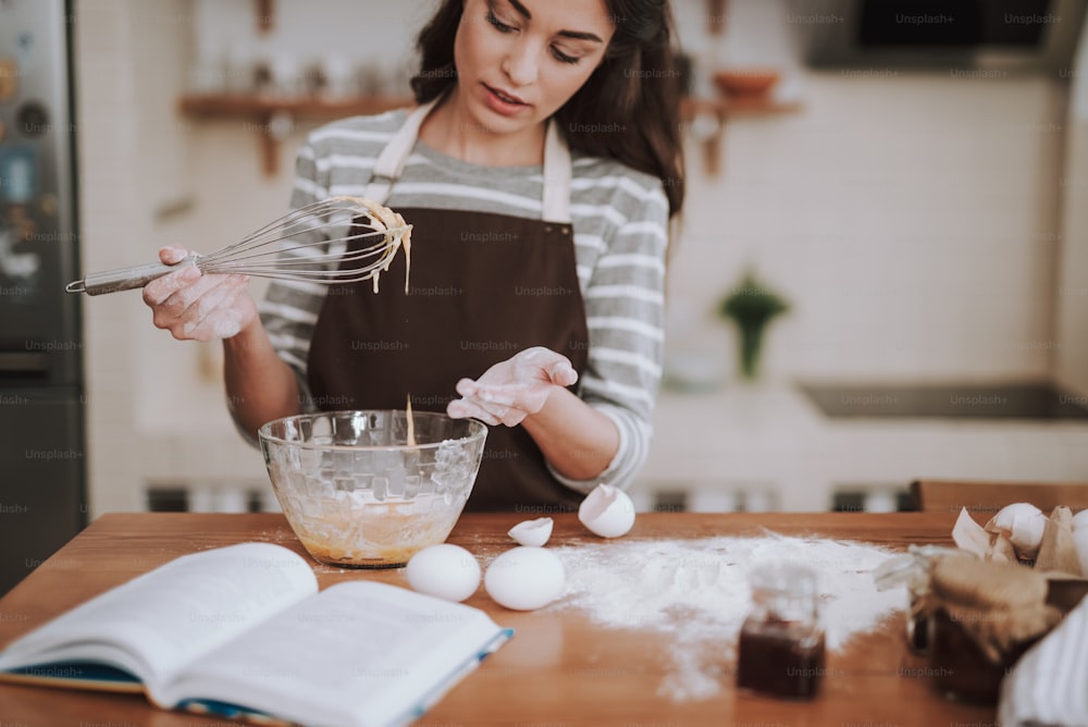 Konzentrieren Sie sich auf Hausfrau, die Bäckerei in der Küche zubereitet. Sie mischt Zutaten mit Schneebesen nach Rezeptbuch