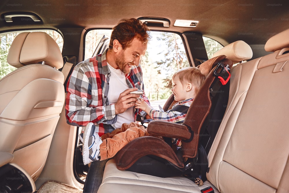 Vater versucht, seinen kleinen Jungen zu unterhalten, der in einem Auto auf einem Sicherheitsstuhl sitzt. Seitenansicht