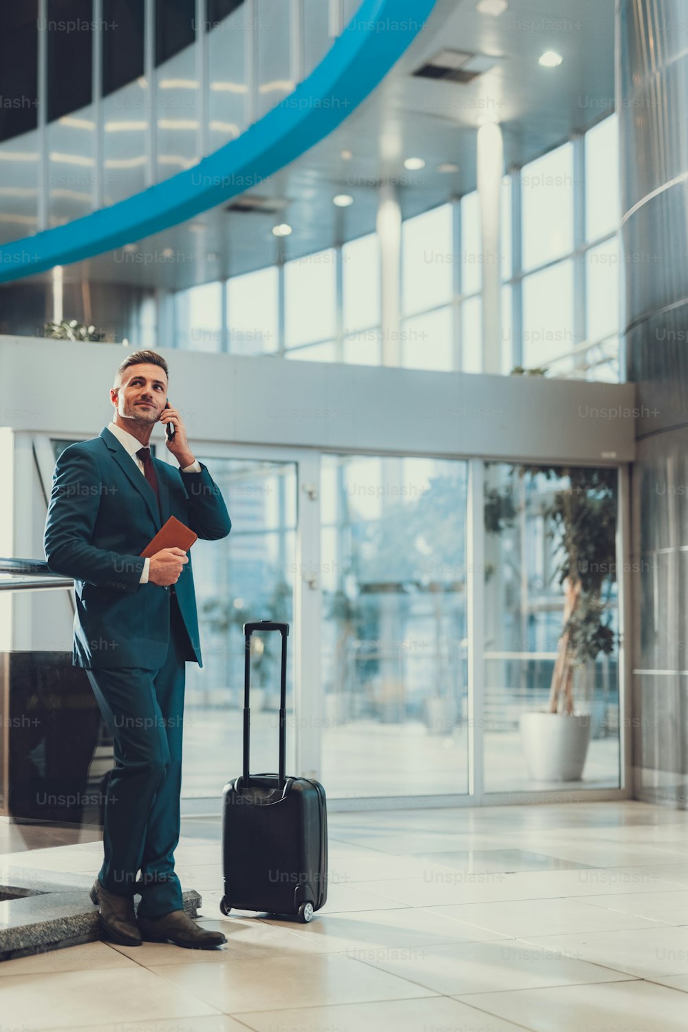 Foto verticale di attraente uomo adulto in abito di classe in piedi vicino alla valigia e in attesa del suo volo. Alza lo sguardo e parla al cellulare