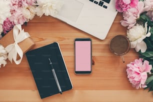 Teléfono elegante con pantalla vacía, computadora portátil, taza de café, cuaderno, regalo con peonías rosas y blancas en la mesa plana con espacio para texto. Concepto freelance. Lugar de trabajo con estilo.