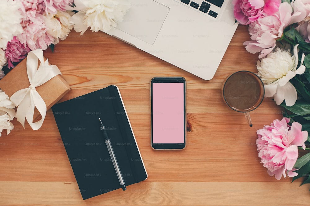 Stylish phone with empty screen, laptop, coffee cup, notebook,gift with pink and white peonies on table flat lay with space for text. Freelance concept. Stylish working place.
