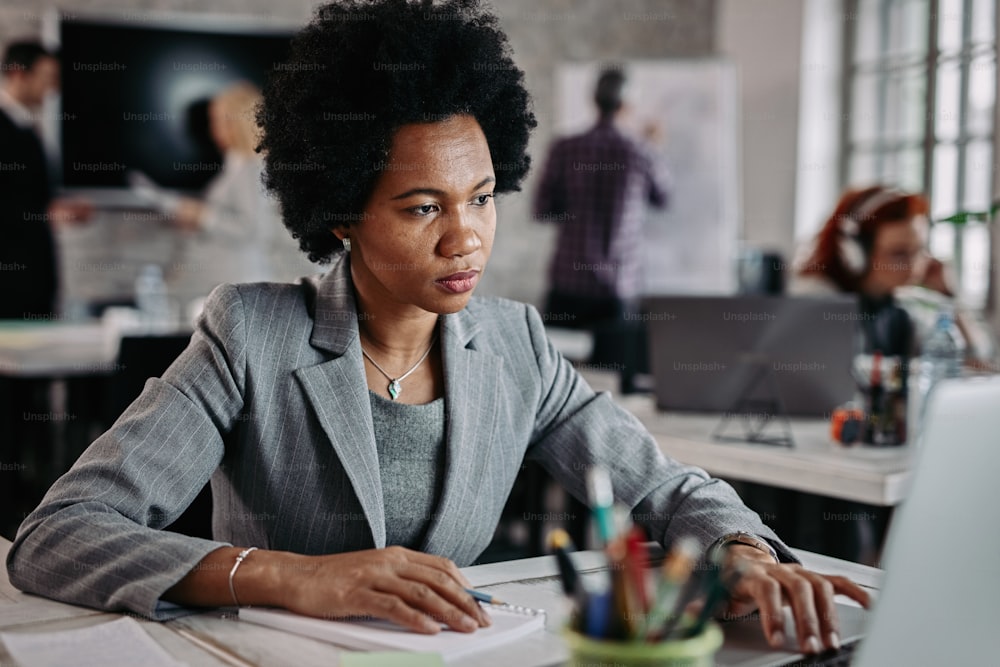 Schwarze Geschäftsfrau im mittleren Erwachsenenalter, die während der Arbeit am Schreibtisch einen Laptop benutzt. Es gibt Leute im Hintergrund.