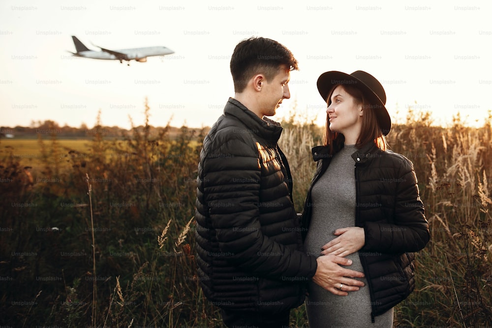 Stylish pregnant couple holding hands on belly in sunny light in autumn park. Happy young parents, mom and dad, hugging baby bump and smiling, enjoying beautiful moment at sunset among grass