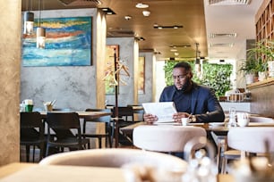 Porträt eines stilvollen jungen afroamerikanischen CEO in dunkelblauer Jacke, der Artikel in einer Zeitung durchblättert, am Cafétisch sitzt und im Café Morgenkaffee trinkt. Modernes Interieur