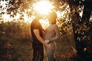 Elegante casal grávida de mãos dadas na barriga e abraçando em luz ensolarada no parque de outono sob a árvore. Jovens pais felizes, mãe e pai, abraçando a barriga do bebê, desfrutando de um belo momento ao pôr do sol
