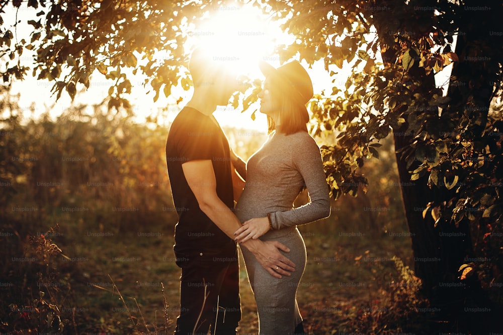 Stylish pregnant couple holding hands on belly and embracing in sunny light in autumn park under tree. Happy young parents, mom and dad, hugging baby bump, enjoying beautiful moment at sunset