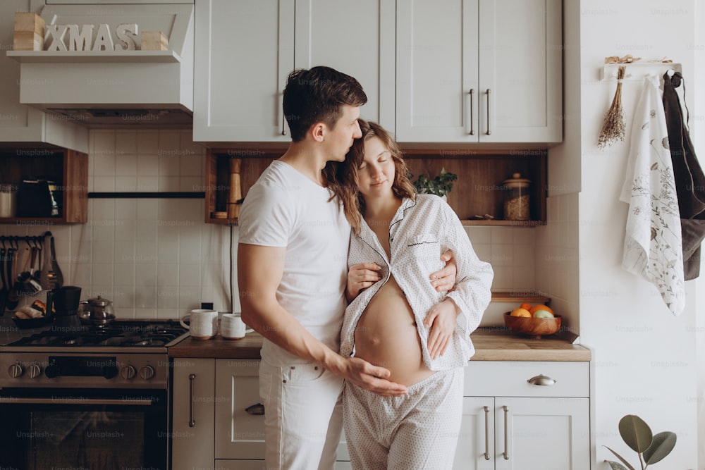 Glückliches junges schwangeres Paar im weißen Schlafanzug, das morgens Bauchbeule in der Küche hält. Stilvolle schwangere Familie, Mama und Papa entspannen sich zu Hause und warten auf Baby. Elternschaft. Süße Momente
