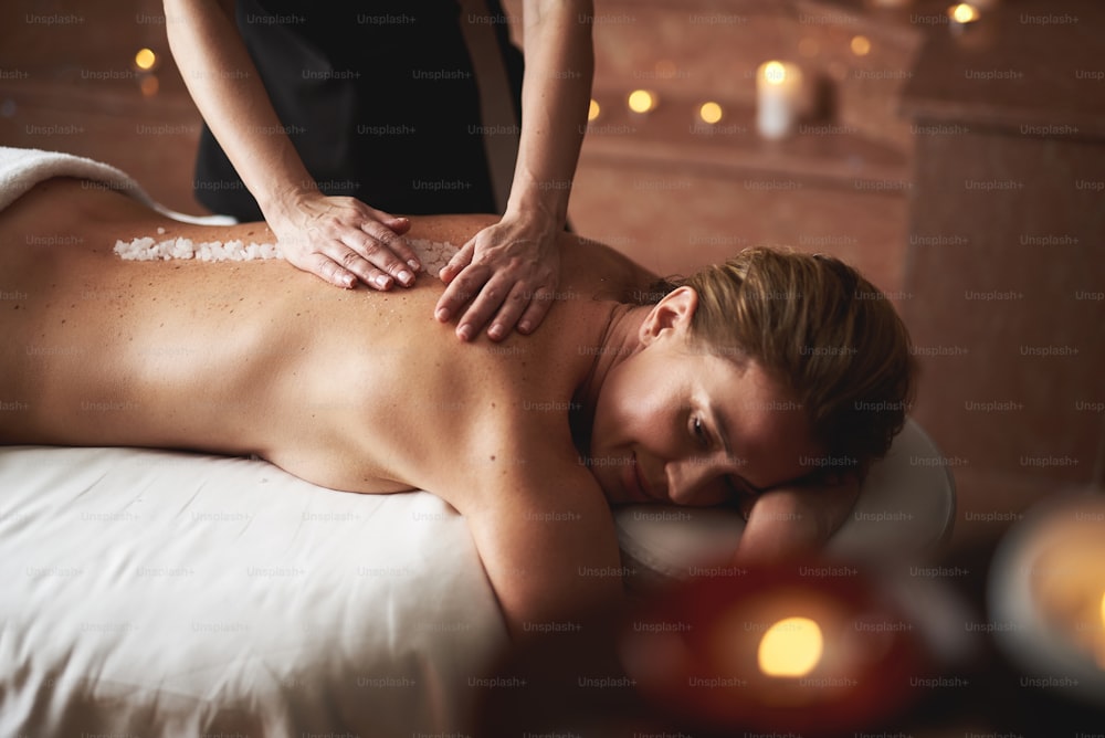 Concept of relaxation and body care. Side view portrait of female therapist applying mineral salt on spine of young lady while making massage in spa health salon