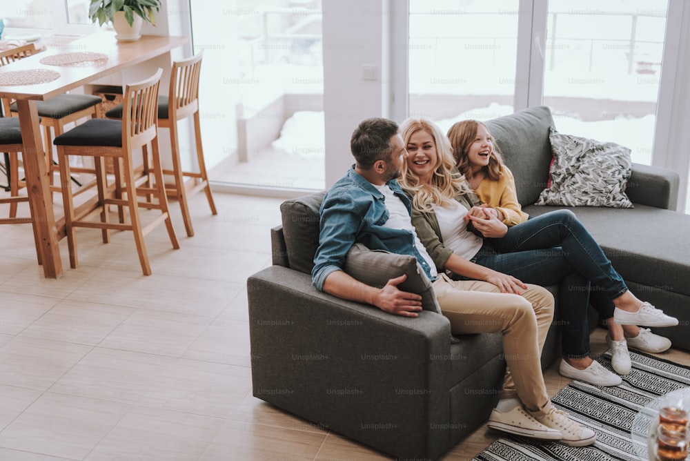 Full length portrait of handsome bearded man sitting on couch with his beautiful wife and daughter. They having fun and laughing