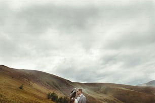 wunderschöne Braut und stilvoller Bräutigam umarmen sich in sonniger Landschaft, Boho-Hochzeitspaar, Luxuszeremonie in den Bergen mit herrlicher Aussicht, Platz für Text
