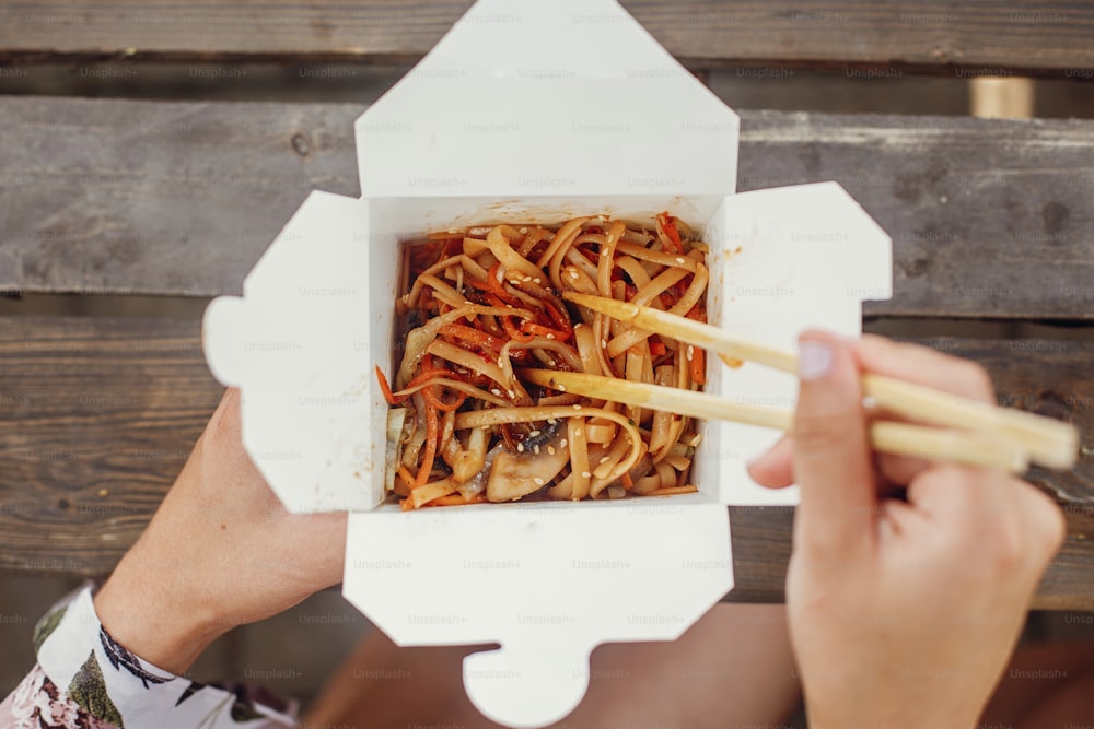 Menina comendo macarrão wok com legumes e frutos do mar na caixa de caixa para ir, com pauzinhos de bambu, close-up. Entrega de comida de cozinha tradicional asiática. Macarrão em caixa de papel takeaway. Comida de rua