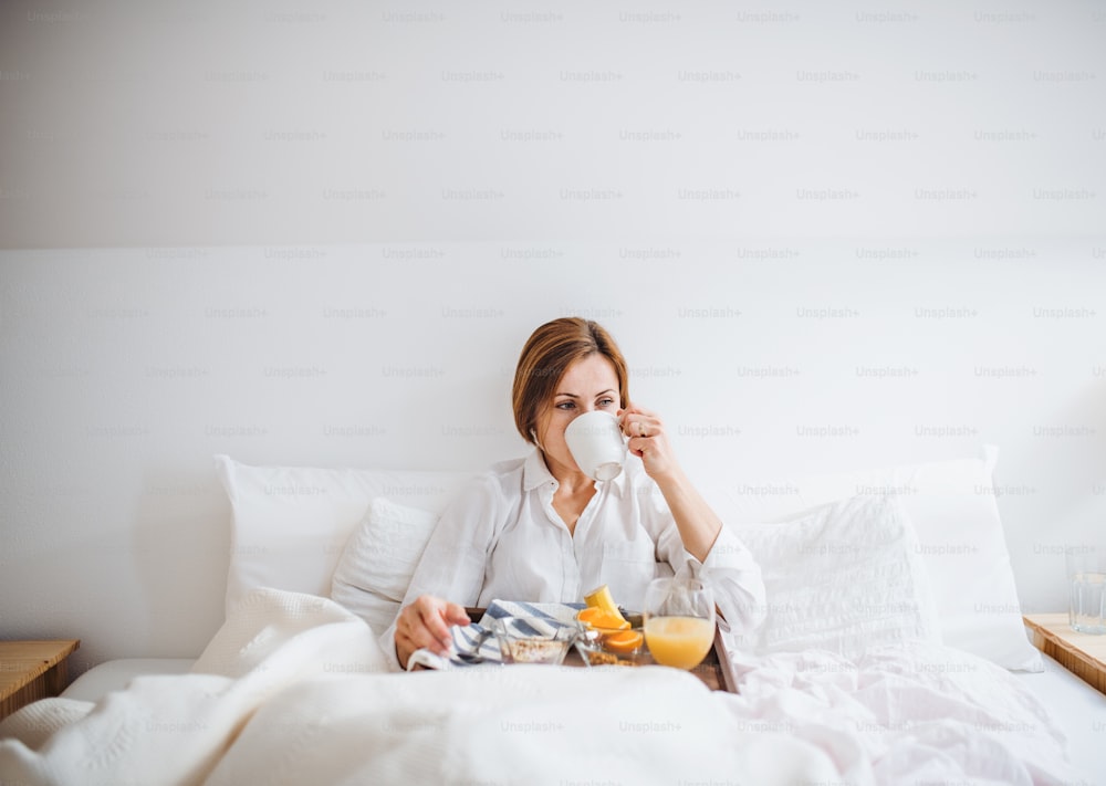 Una vista frontale di una giovane donna sdraiata a letto con caffè e colazione al mattino in una camera da letto.