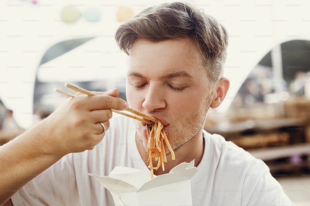 Stylish hipster man eating delicious wok noodles with vegetables from carton box with bamboo chopsticks. Asian Street food festival. Guy tasting and eating thai noodles in takeaway paper box