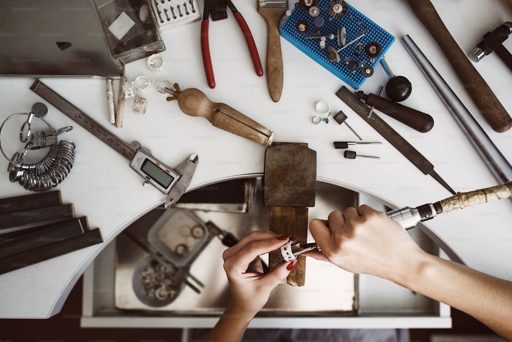 Creative chaos. Top view of jeweler's workbench with different tools for making jewelry. Female jeweler's hands polishing a silver ring with grinding machine. Jewelry making process. Jewelry equipment. Working process. Jewelry manufacturing concept.